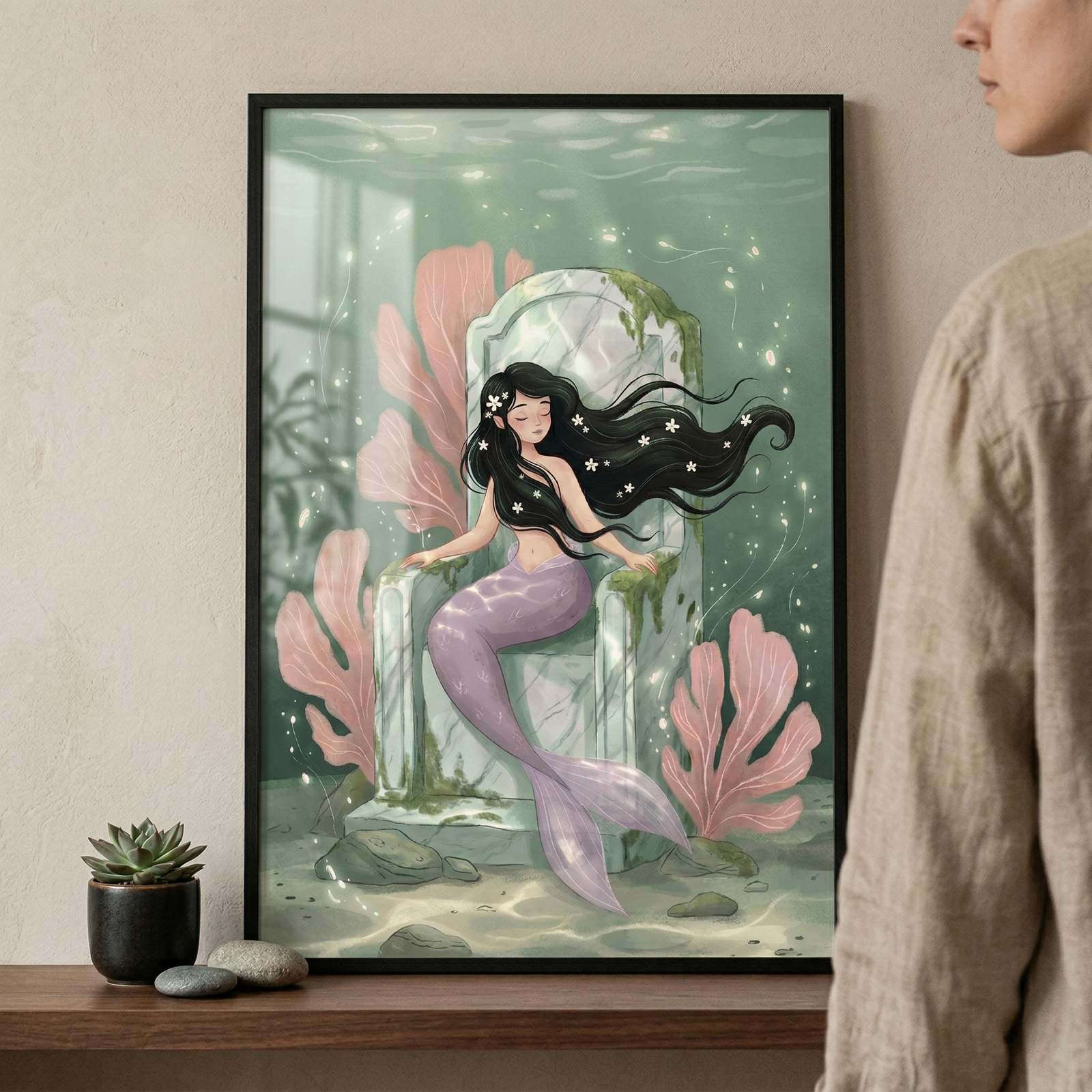 A framed illustration shows a mermaid with long, flowing black hair sitting on a stone throne underwater. Her tail is light purple, and small white flowers are tucked into her hair. She is surrounded by large pink coral plants and glowing bubbles in a soft teal sea. Gentle light filters from the surface above. The frame sits on a wooden shelf next to a small potted succulent and smooth stones. 