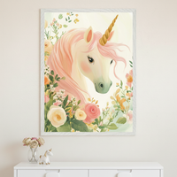 A person in a beige sweater holds up a framed illustration of a unicorn with a golden horn, soft pink mane, and gentle expression, surrounded by pastel-colored flowers including roses and greenery. The whimsical artwork has a dreamy, fairytale-like style, and the person’s face is hidden behind the frame.