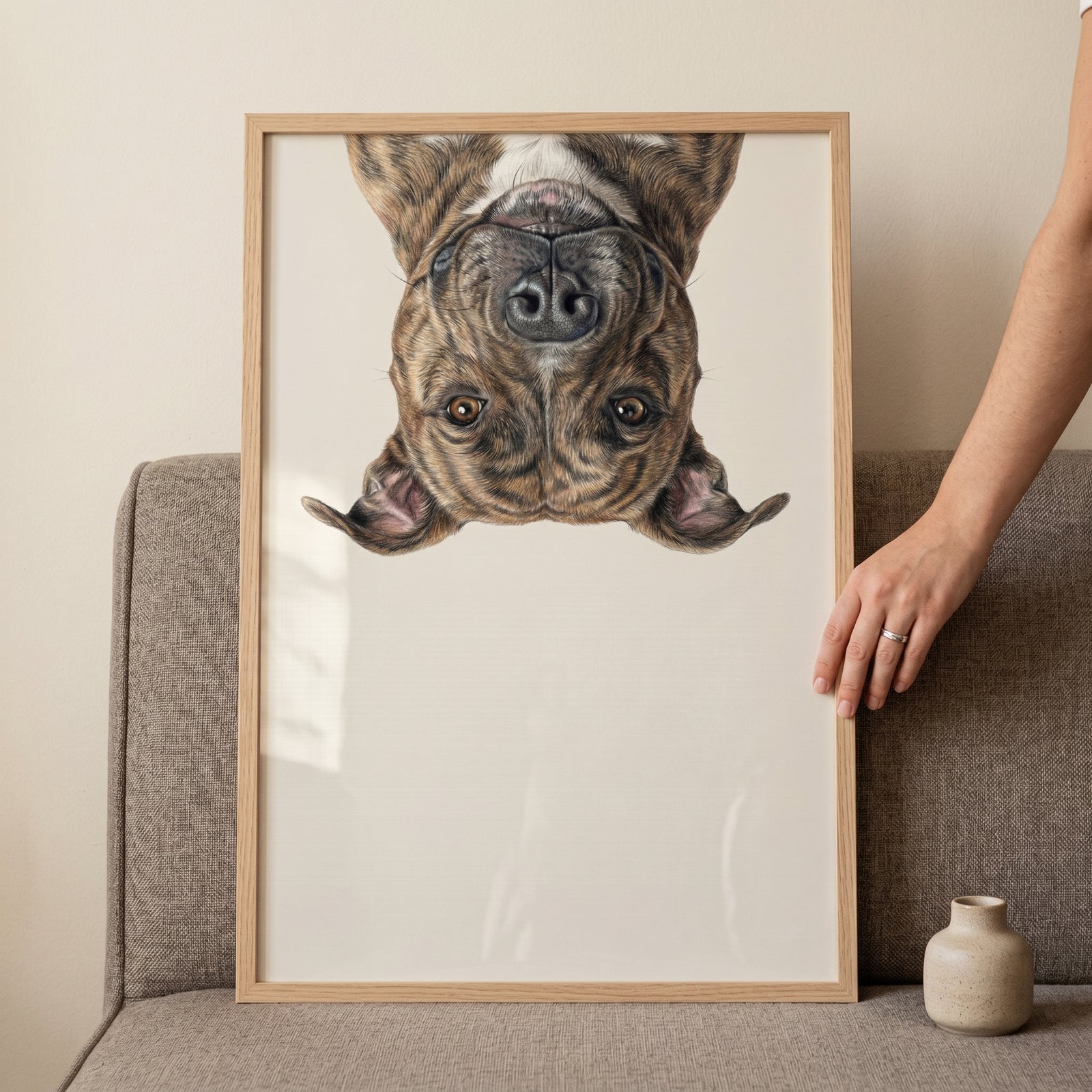 Framed upside down brindle dog wall art held by a hand against a neutral wall and grey fabric couch.