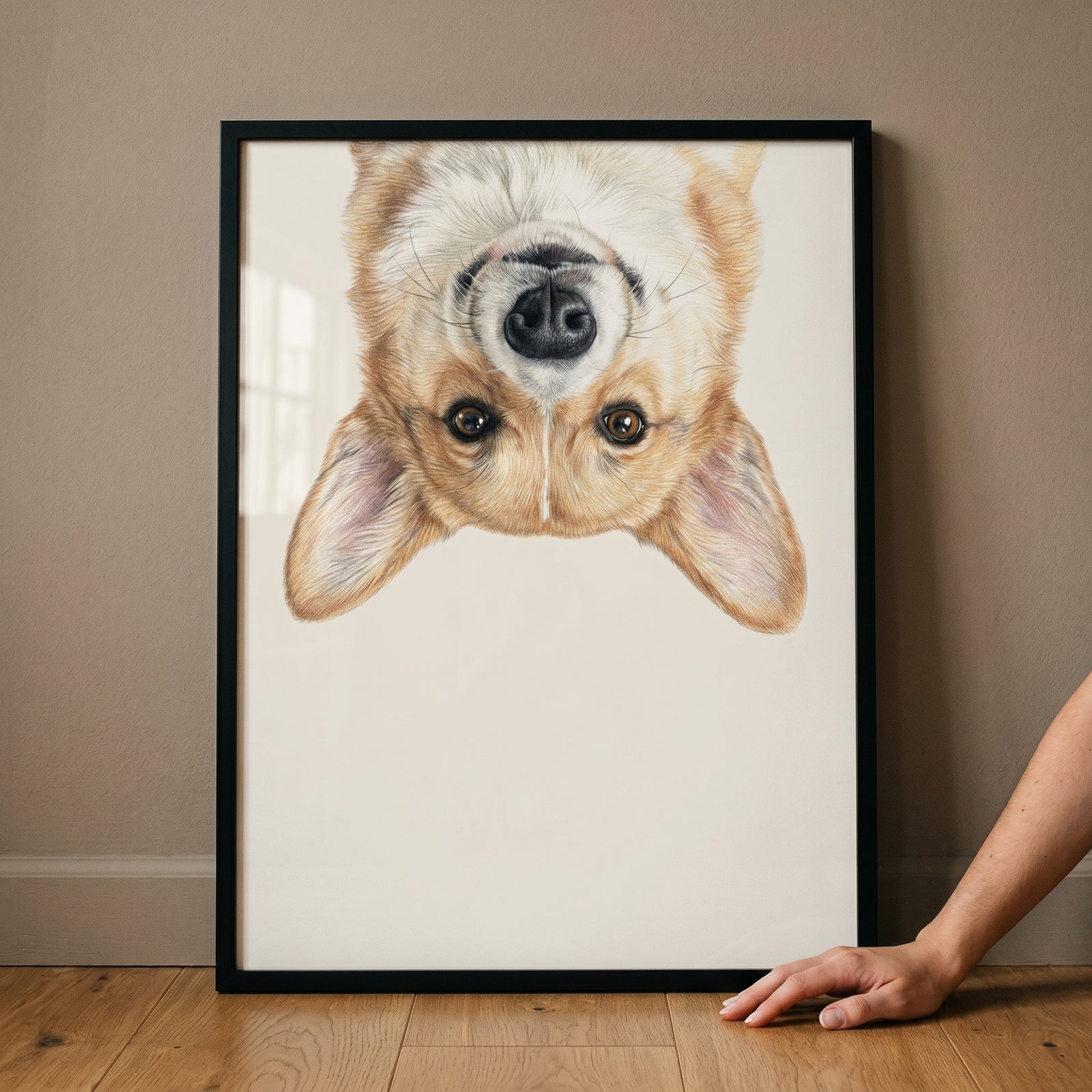 Upside down tan corgi wall art in a black frame leans against a wall while a hand rests on the floor nearby.