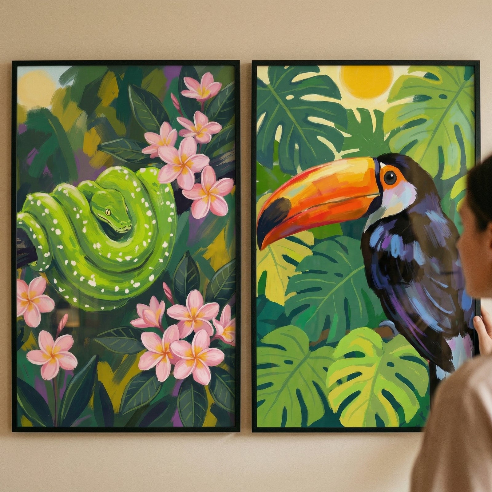 Two framed jungle themed paintings hang on a wall while a person looks at them from the side. One shows a bright green snake coiled among dark leaves and pink flowers while the other shows a toucan with a large orange beak perched among tropical leaves under a yellow sun.