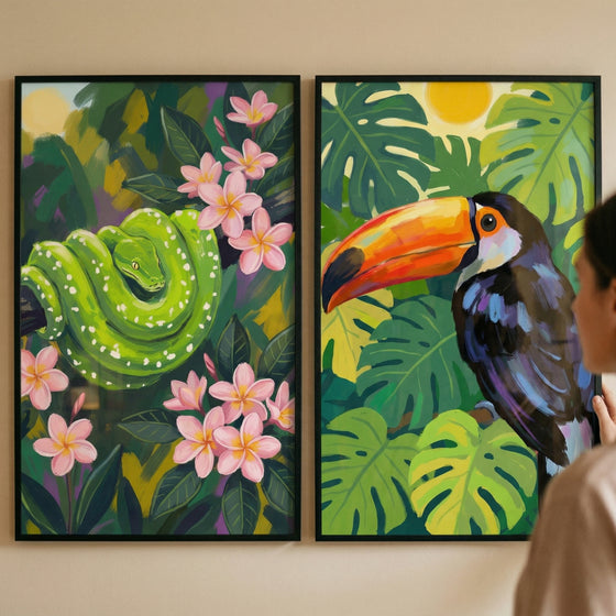 Two framed jungle themed paintings hang on a wall while a person looks at them from the side. One shows a bright green snake coiled among dark leaves and pink flowers while the other shows a toucan with a large orange beak perched among tropical leaves under a yellow sun.