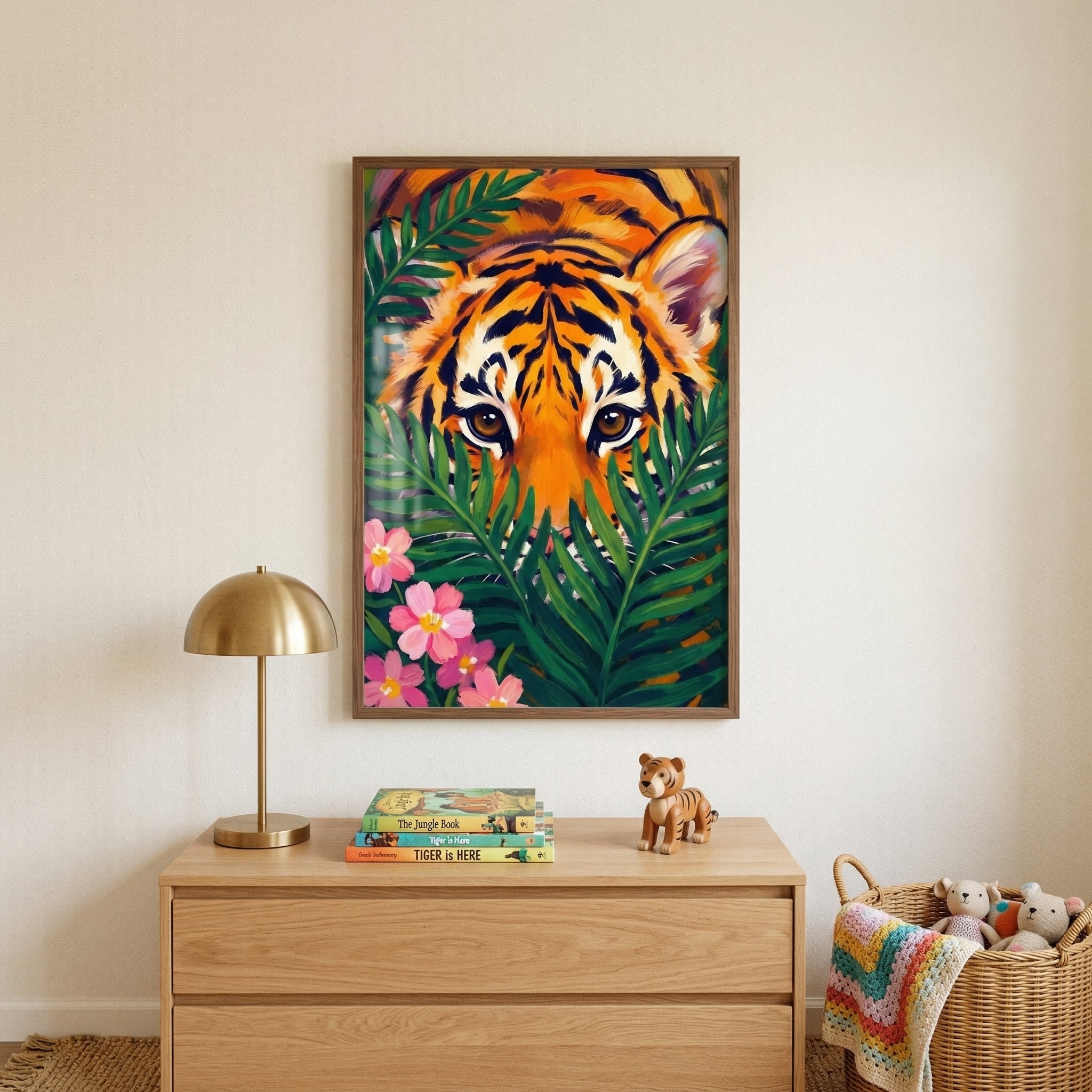 A vertical image shows a light-filled child's room with neutral beige walls. Centered on the wall is a framed illustration of a tiger's face, rendered with bold orange and black stripes and piercing eyes, peering through large green tropical leaves. Several pink blossoms are visible in the lower-left corner of the artwork. 
