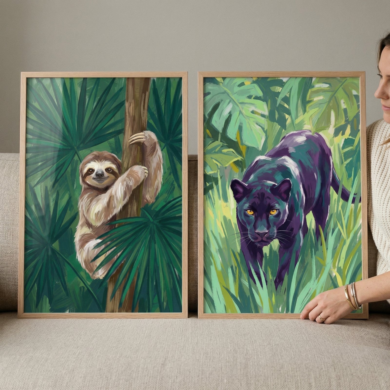 A person holds two framed illustrations showcasing stylized jungle animals. The frame on the left features a three-toed sloth clinging to a vertical tree trunk, set against a background of large, lush green palm fronds. The frame on the right displays a sleek black panther with glowing yellow eyes, depicted walking through dense tall grass and tropical foliage. Both artworks are presented in light-toned wooden frames and utilize a vibrant color palette, including deep greens, purples, and earthy browns.