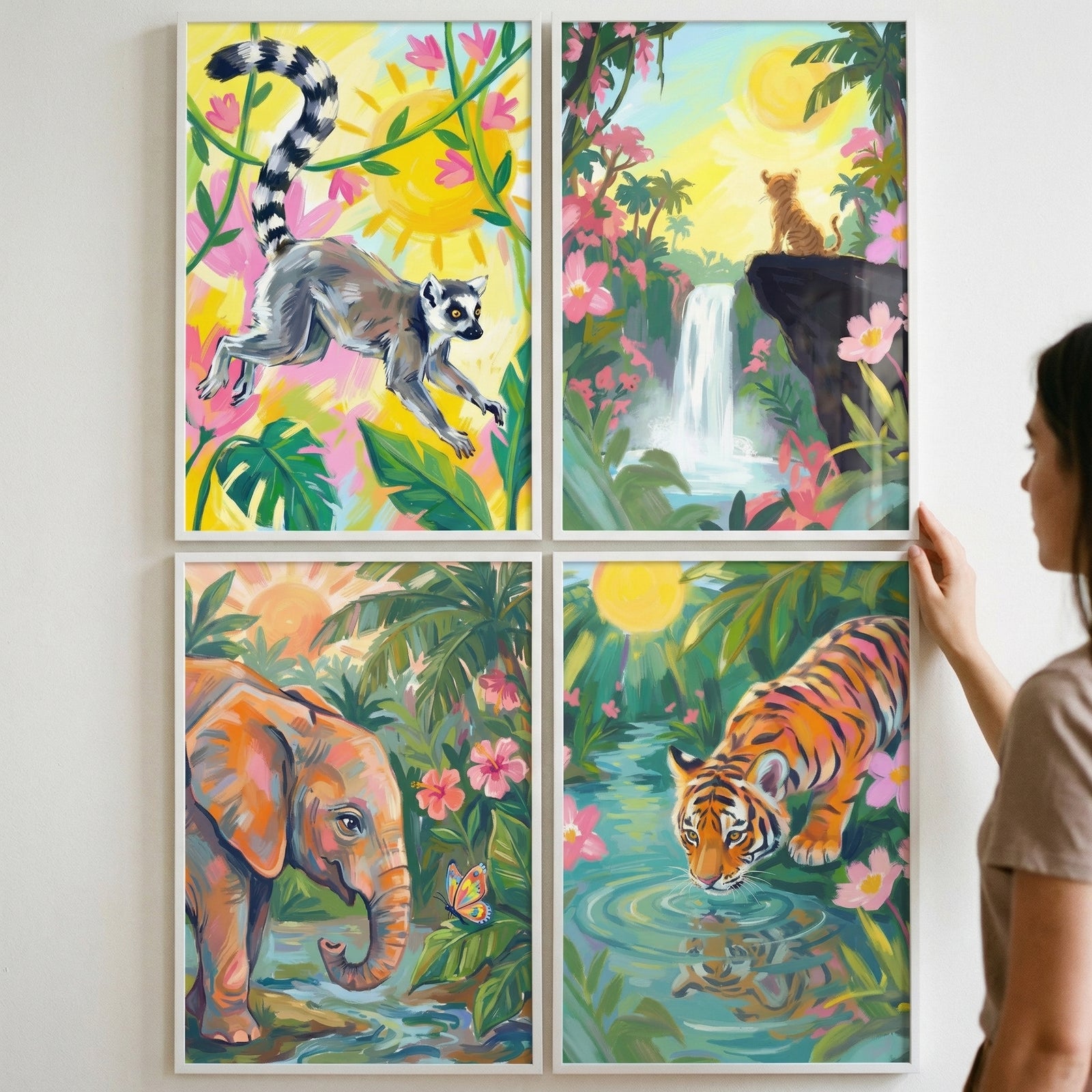 Four framed jungle themed paintings arranged in a grid on a wall while a person adjusts the top right frame. The artwork features a lemur leaping among bright flowers, a tiger sitting above a waterfall at sunrise, an elephant standing in a lush river scene with a butterfly, and a tiger drinking from water surrounded by tropical plants.