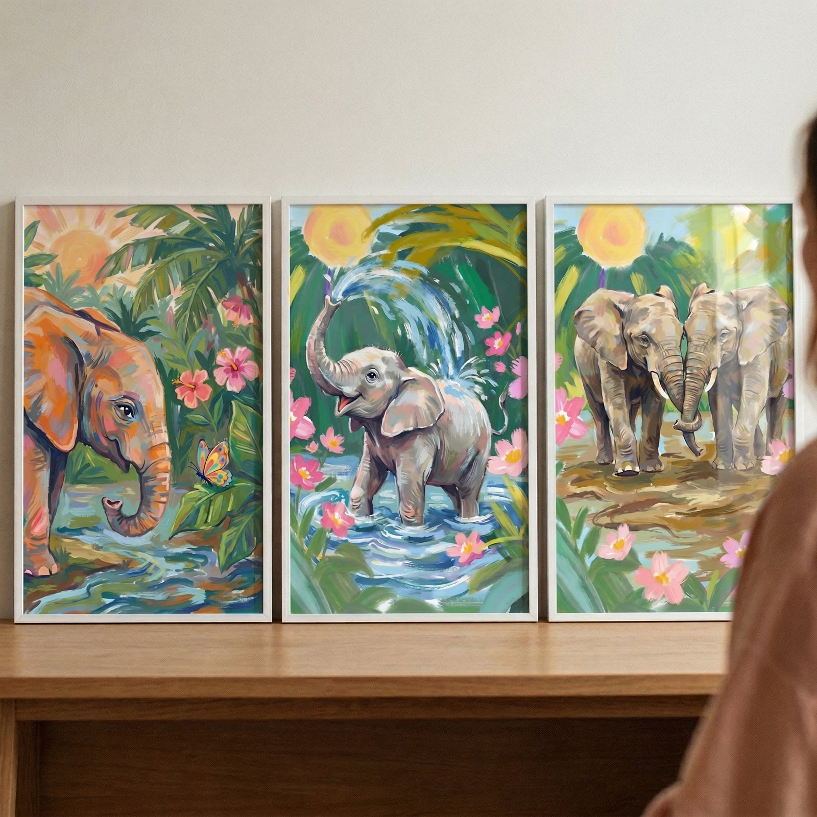 Three framed paintings of elephants in a colorful jungle setting are displayed side by side on a wooden surface while a person stands nearby. The artwork shows an elephant by a stream with a butterfly, a baby elephant splashing water with its trunk among pink flowers, and two elephants standing together in a sunlit tropical scene.