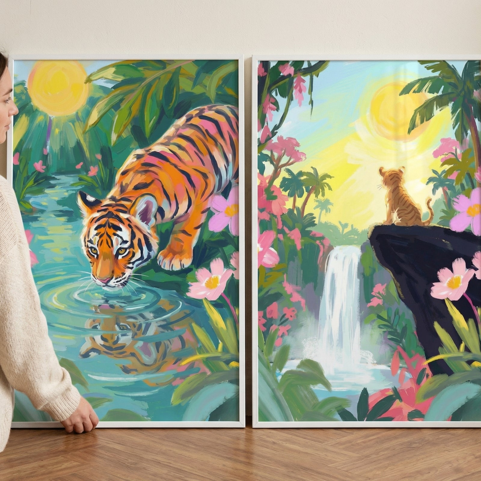 Two framed jungle paintings lean against a wall while a person adjusts the left frame. One shows a tiger drinking from a calm river surrounded by green leaves and pink flowers while the other shows a tiger sitting on a cliff overlooking a waterfall and tropical landscape under a bright sun.
