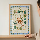 A framed illustration shows a cheerful brown cartoon monkey swinging from a green vine. The smiling monkey is surrounded by stylized jungle leaves and small colorful flowers on a cream background. A light blue border with green stripes surrounds the artwork inside a light wood frame. The frame sits on a wooden shelf against a plain wall, with a person's hand visible on the right side adjusting its position.