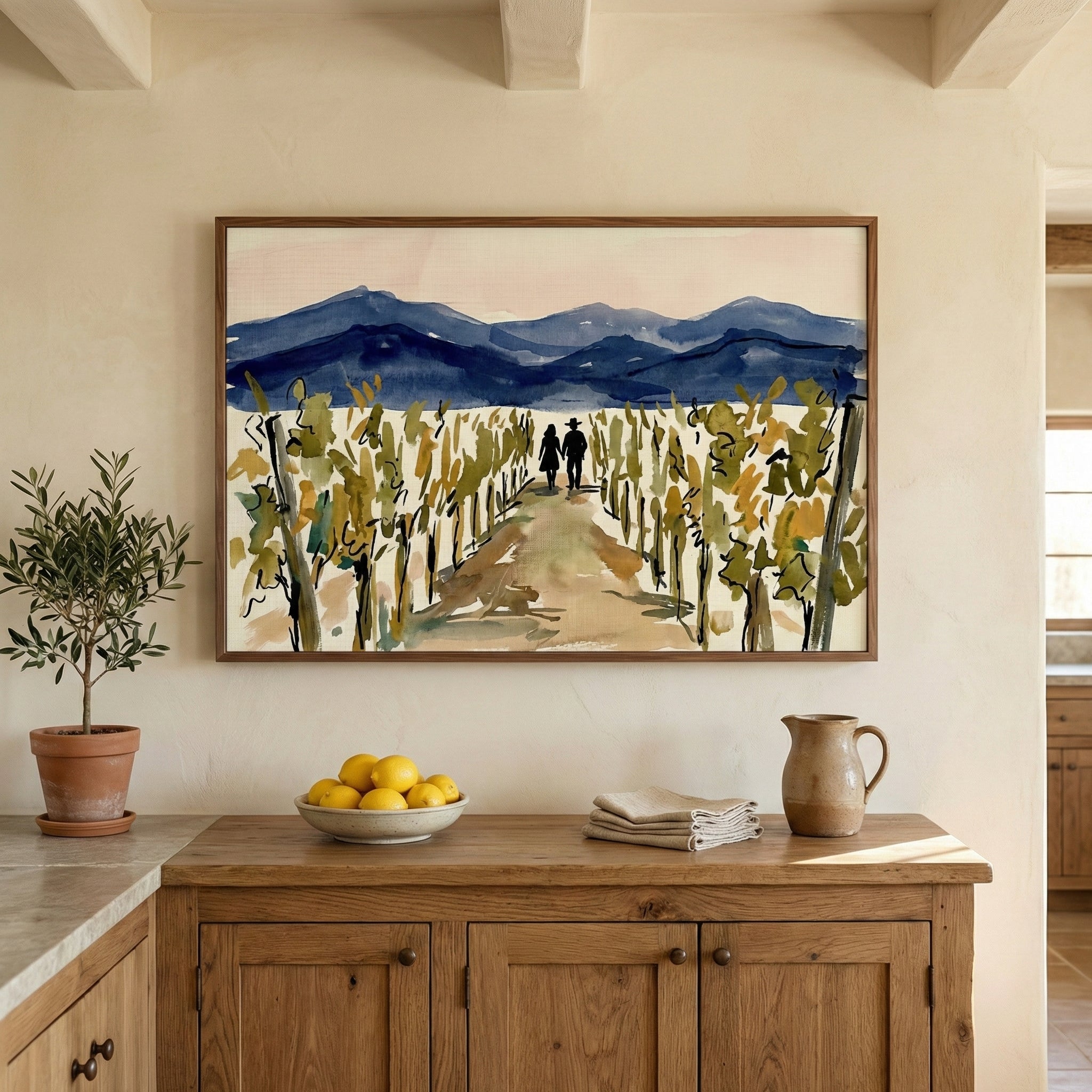 A minimalist interior with a large framed landscape illustration hanging on a neutral-colored wall. The artwork depicts two people walking down a path through a vineyard with blue mountains in the distance. Below the frame, a rustic wooden cabinet holds a small olive tree in a terracotta pot, a bowl of lemons, a stack of white napkins, and a tan ceramic pitcher. The room features exposed wooden ceiling beams and a warm, earthy color palette.