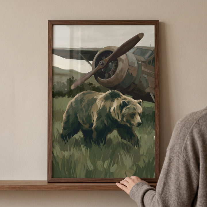 Framed vintage airplane brown bear wall art on a shelf shows a bear in tall grass in front of an old propeller aircraft.
