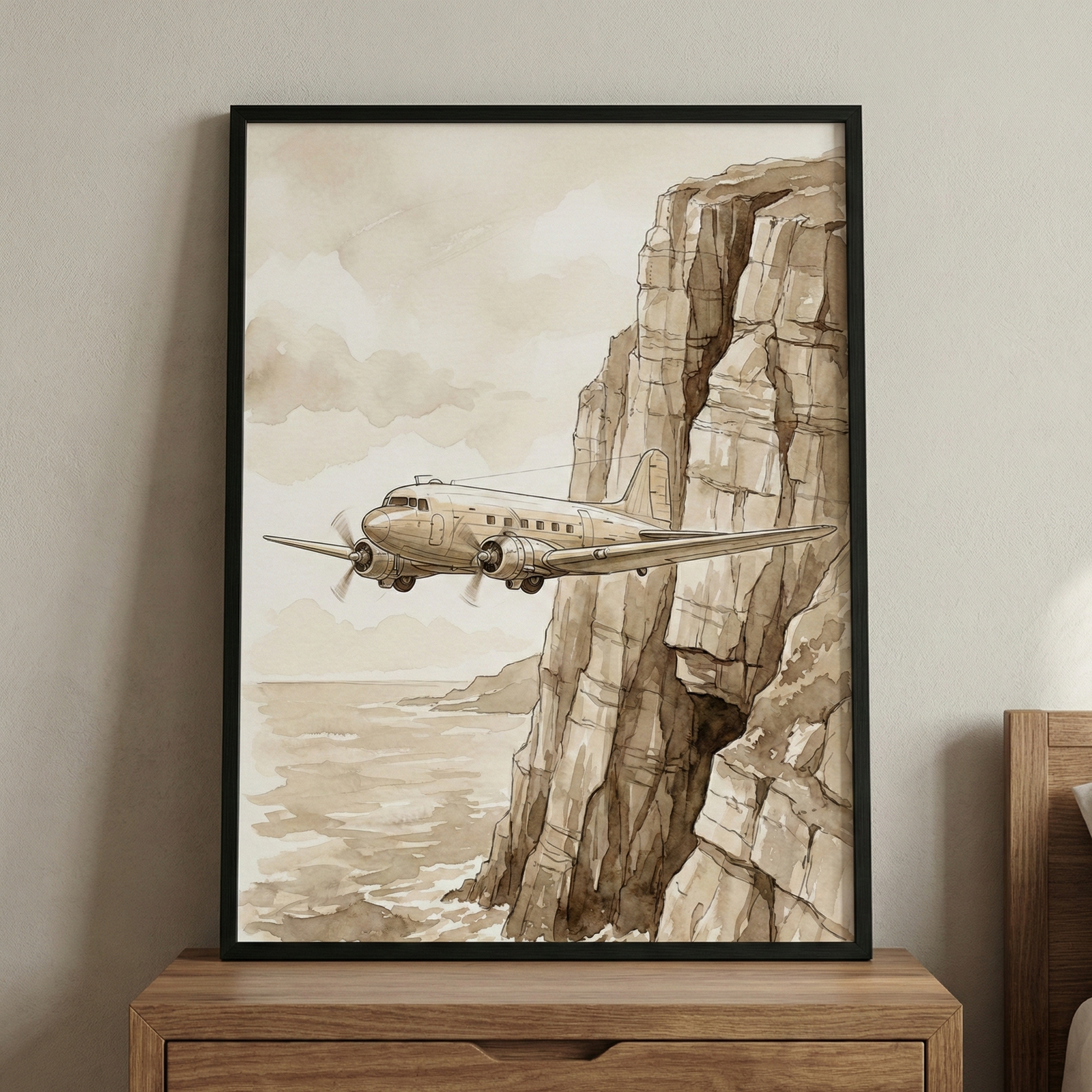 A framed watercolor-style illustration depicts a vintage twin-engine propeller aircraft soaring alongside a towering, rugged cliff. The scene is rendered in a monochromatic sepia and tan color palette, giving it a nostalgic feel. Below the steep rock face, the ocean waves are suggested with soft brushstrokes, and the sky is filled with pale, hazy clouds. The artwork is showcased in a simple black frame, resting on a light-colored wooden cabinet against a neutral-toned wall.