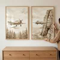 Two framed sepia watercolor illustrations are hung on a white wall above a wooden dresser. A person's hands are visible as they adjust the frame on the right. Both artworks feature vintage aircraft; the one on the left shows a single-propeller plane flying over a dense pine forest, while the one on the right depicts a twin-engine plane soaring past a towering coastal cliff. The frames are made of light wood, matching the dresser below, which has four drawers with simple round knobs.