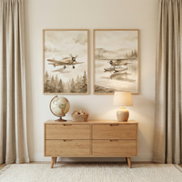 A nursery setup features a light wooden dresser against a cream wall. On top sits a globe, a small woven basket, and a glowing table lamp. Above the dresser, two framed watercolor-style illustrations hang side-by-side. The left shows a vintage airplane soaring over pine trees; the right depicts a seaplane on a misty lake. Long, light beige curtains frame the scene, which is set on a white textured rug. The overall aesthetic is warm, neutral, and cozy.