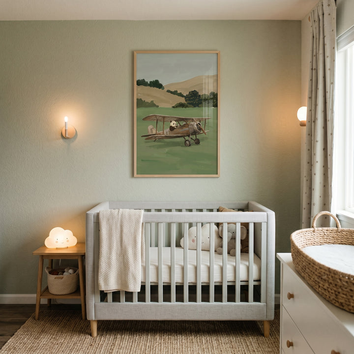 A nursery with a grey crib, cloud lamp, and vintage biplane pilot panda wall art on sage green walls.