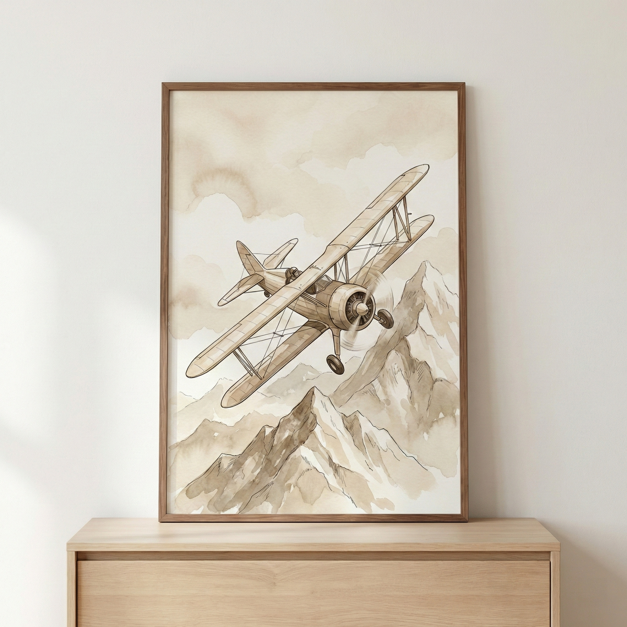 A vertical framed illustration of a vintage biplane soaring over a rugged mountain range. The artwork is rendered in a monochromatic sepia and beige color palette with soft, washed-out textures. The plane is positioned at a diagonal angle above the jagged peaks, set against a cloudy sky. The framed print is displayed on a light wood dresser against a neutral wall.