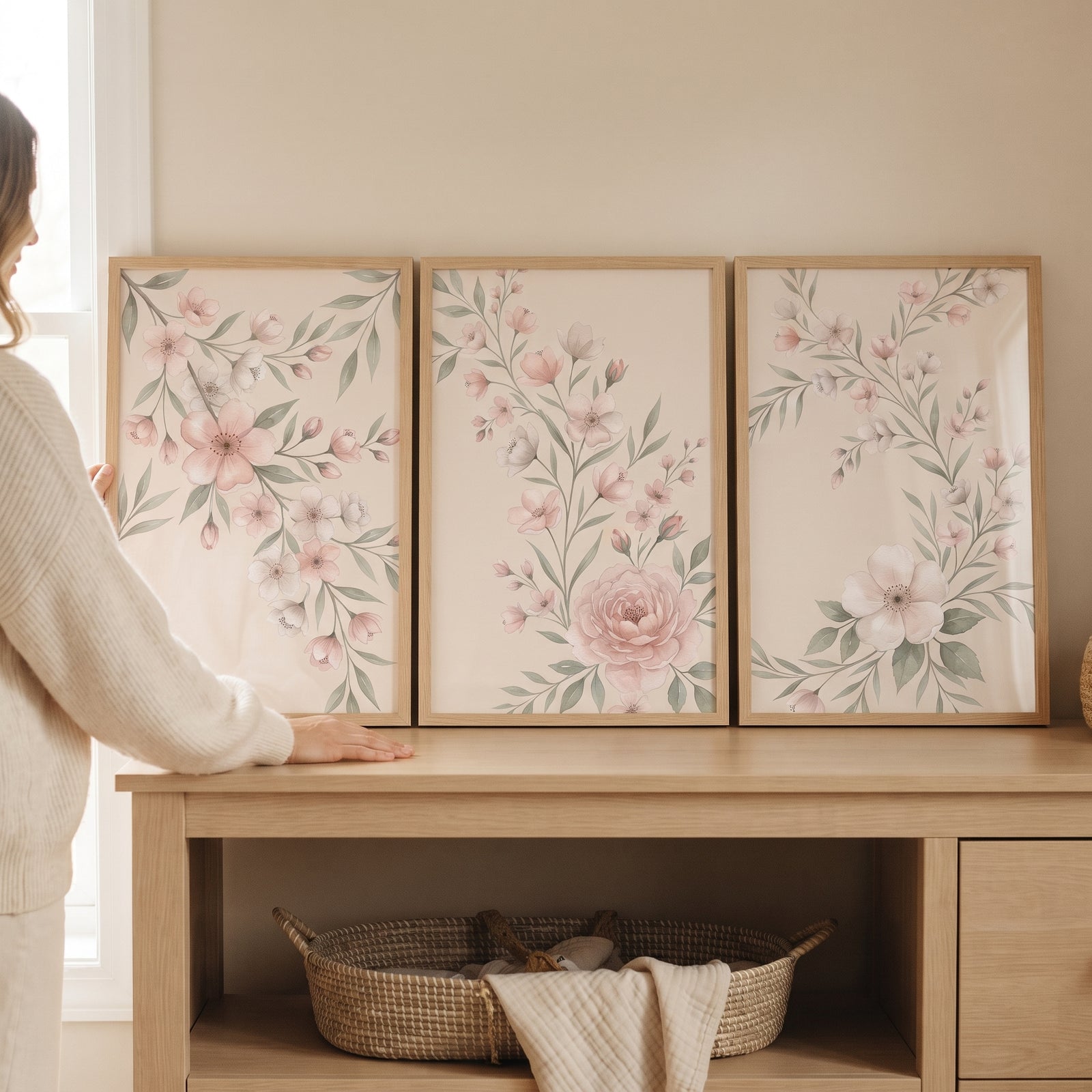 Vintage blush pink floral wall art set of 3 on a wooden table as a person in a sweater adjusts the frames.