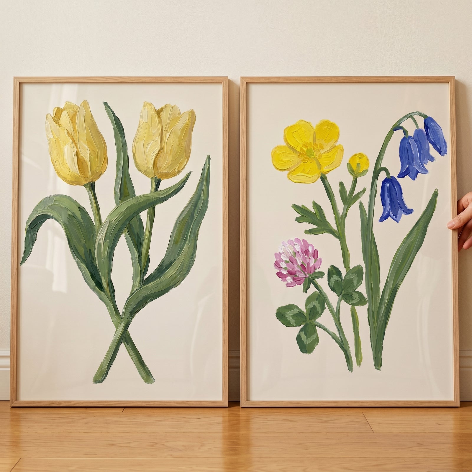 Two framed botanical paintings displayed side by side on a wooden floor featuring yellow tulips on the left and a mix of yellow, blue, and pink flowers on the right, with a hand gently holding the edge of one frame.