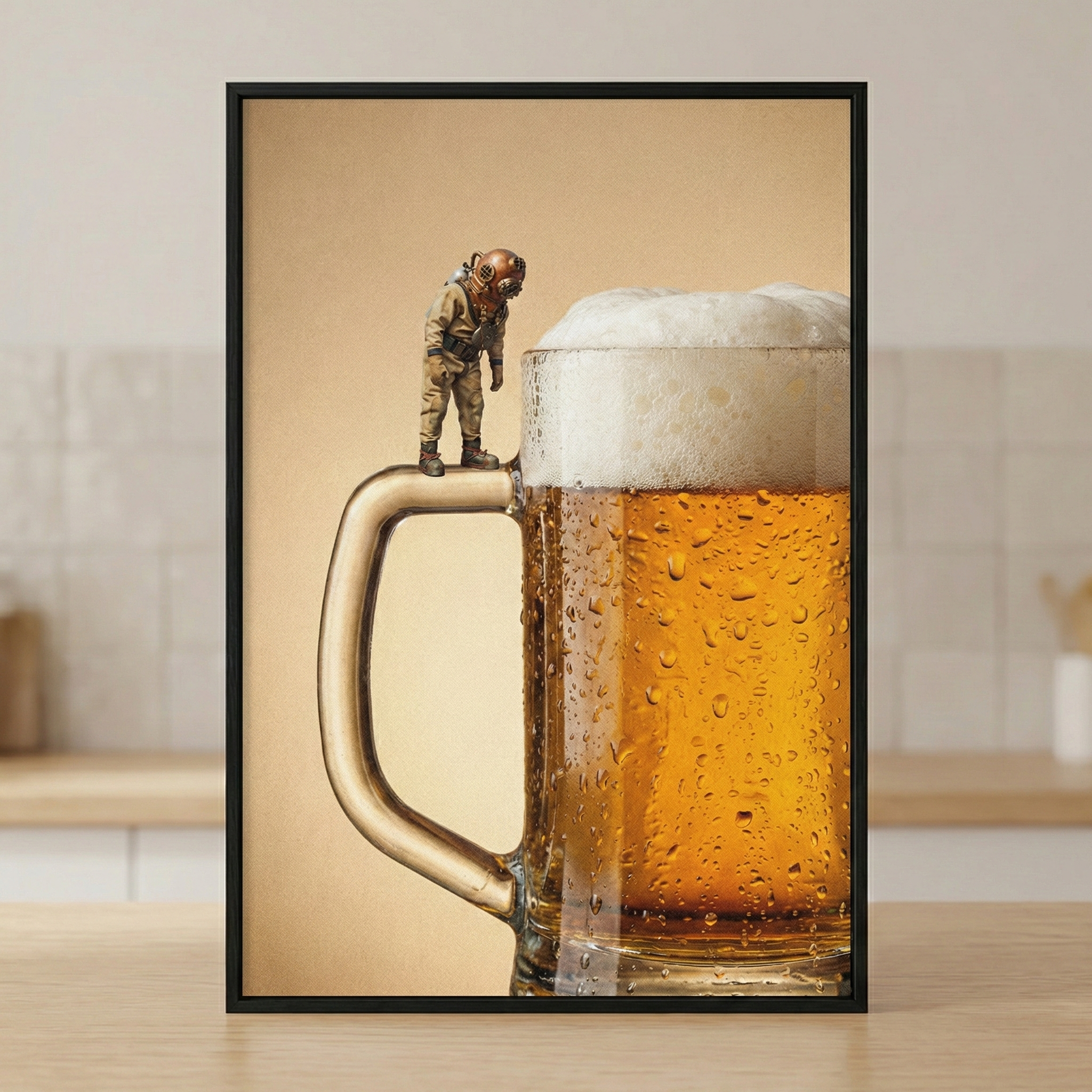 A framed image depicts a large glass mug of beer with a thick, frothy head of foam. Tiny droplets of condensation cling to the outside of the glass. On top of the mug's sturdy handle, a miniature figure of a vintage deep-sea diver in a brass-colored helmet and bulky suit stands looking down into the foam. The background is a solid, neutral tan color, and the entire scene is enclosed in a slim black frame, resting on a wooden surface.