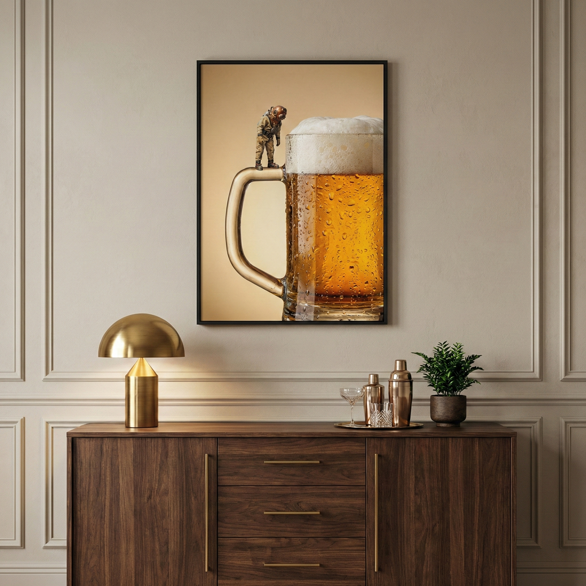A framed piece of wall art depicting a surreal scene of a miniature vintage deep-sea diver standing on the handle of a large, frothy mug of beer. The mug is filled with a golden liquid and covered in condensation. The art is centered on a beige wall with decorative molding above a dark wood sideboard. On the sideboard, a brass mushroom-shaped lamp sits to the left, and a small potted plant and gold-toned cocktail shakers are arranged on the right.