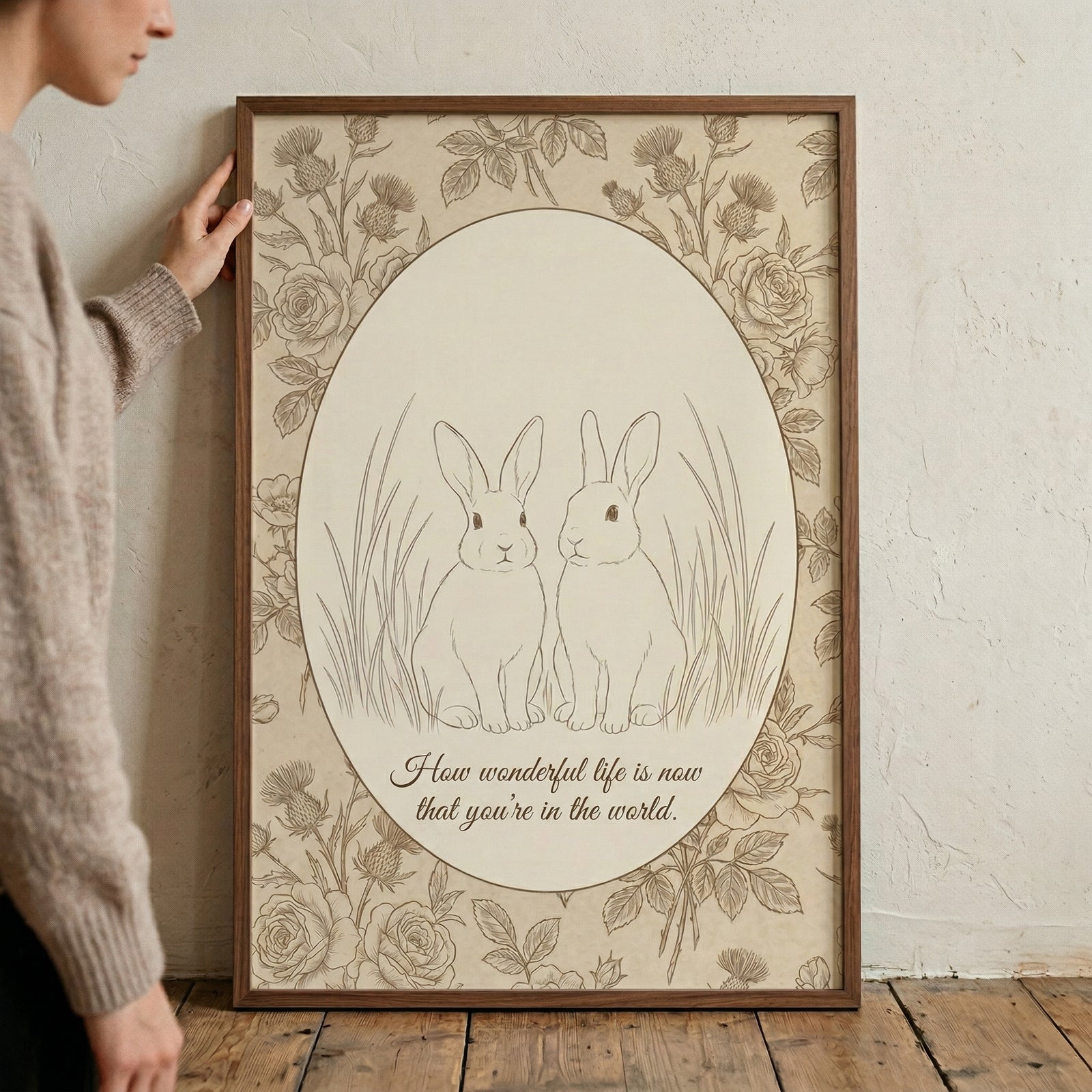 Framed vintage style illustration of two rabbits sitting side by side within an oval border, surrounded by detailed floral designs. The text "How wonderful life is now that you're in the world." appears below them, creating a sentimental and gentle nursery themed print.