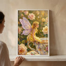 An impressionistic artwork is displayed in a simple white frame, resting on a clean white shelf. The piece features a small, delicate fairy with translucent, purple-tinted wings sitting gracefully on the rim of an oversized teacup. The fairy has long, golden-blonde hair and is dressed in a soft yellow gown. 