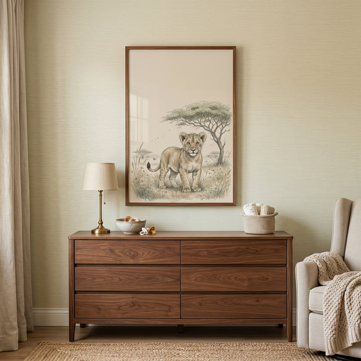 Framed vintage lion cub wall art hangs above a wood dresser in a neutral nursery with a lamp and armchair.