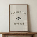 Framed minimalist print with the words "LONG LIVE Boyhood" and a small cowboy hat illustration centered between the text, displayed on a wooden surface. The simple neutral design gives a playful western themed touch to a nursery or kids room.