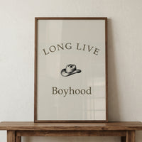 Framed minimalist print with the words "LONG LIVE Boyhood" and a small cowboy hat illustration centered between the text, displayed on a wooden surface. The simple neutral design gives a playful western themed touch to a nursery or kids room.