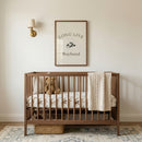 Wooden crib styled with a teddy bear and knit blanket sits beneath a framed print that reads "LONG LIVE Boyhood" with a small cowboy hat illustration. The neutral nursery features soft textures and warm tones, creating a calm and cozy space.