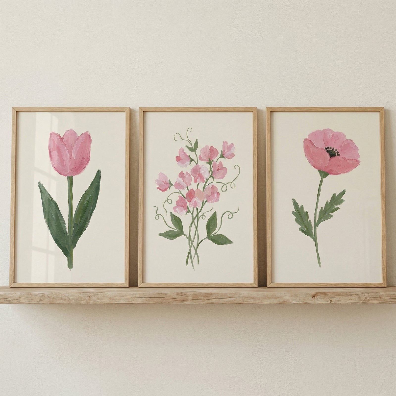 Three framed botanical paintings sit on a wooden shelf against a neutral wall. The artwork features a pink tulip on the left, a cluster of small pink flowers in the center, and a single pink poppy on the right, creating a soft and cohesive floral decor display.