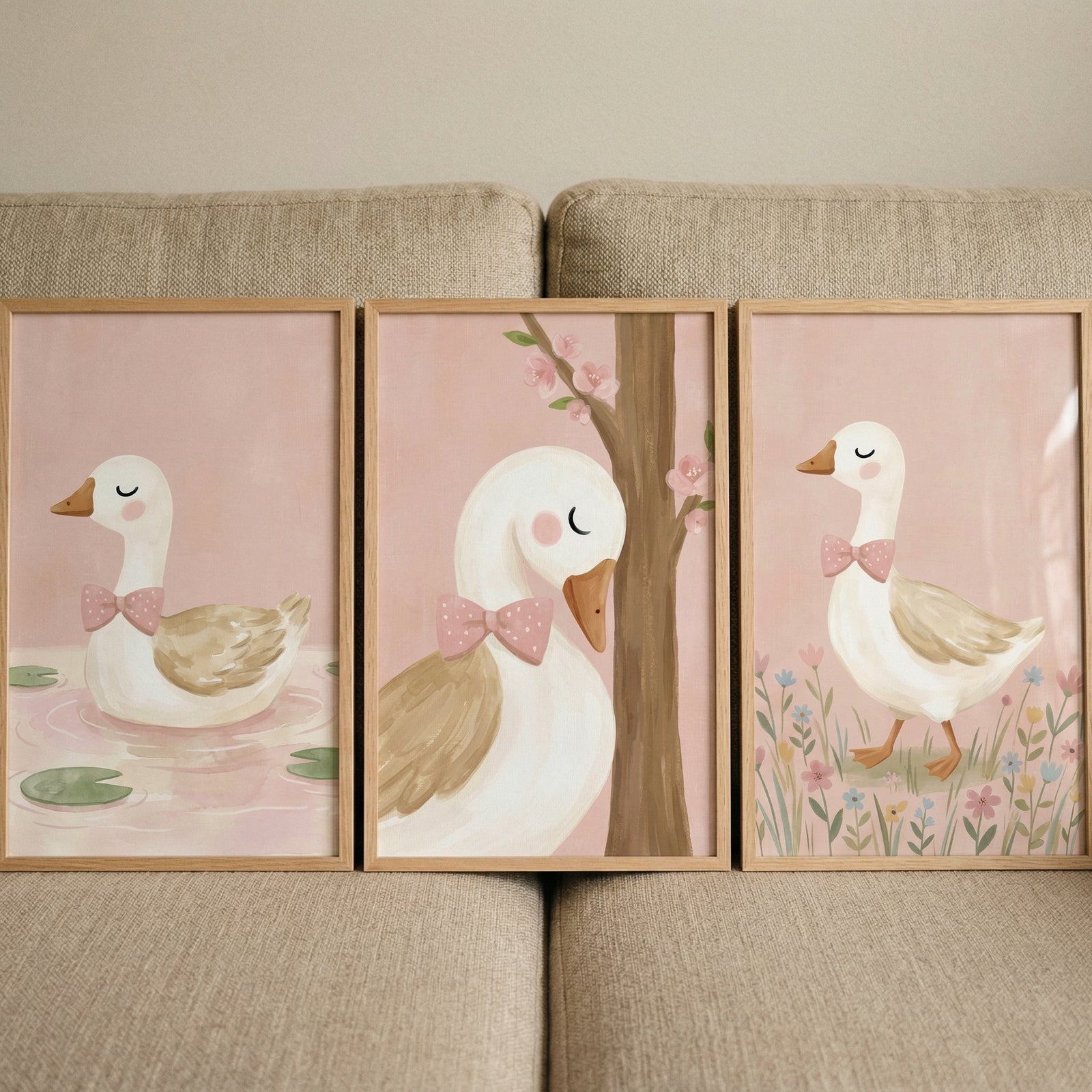Three framed pastel duck illustrations are displayed side by side on a neutral sofa, each duck wearing a pink bow tie against a soft blush background. One duck floats on water with lily pads, one rests beside a flowering tree, and one stands among small flowers, creating a gentle and cohesive nursery art set.