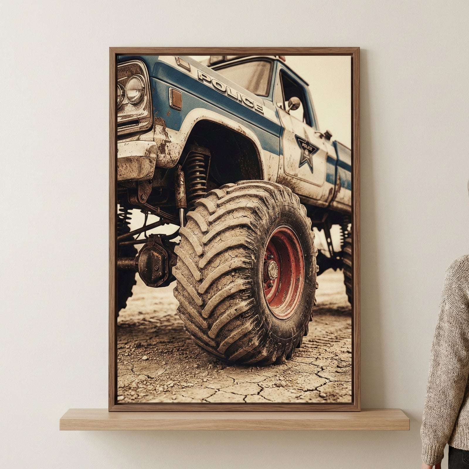 A vertical framed artwork of a vintage-style monster truck with massive, treaded tires and a high suspension system. The weathered utility vehicle features a blue and white color scheme with generic law enforcement markings, including the word "POLICE" and a star emblem on its side. The truck is shown parked on a parched, cracked earth surface. The piece is displayed in a thin wooden frame hanging on a neutral-toned wall above a matching wooden shelf. A person's shoulder wearing a textured beige sweater is 