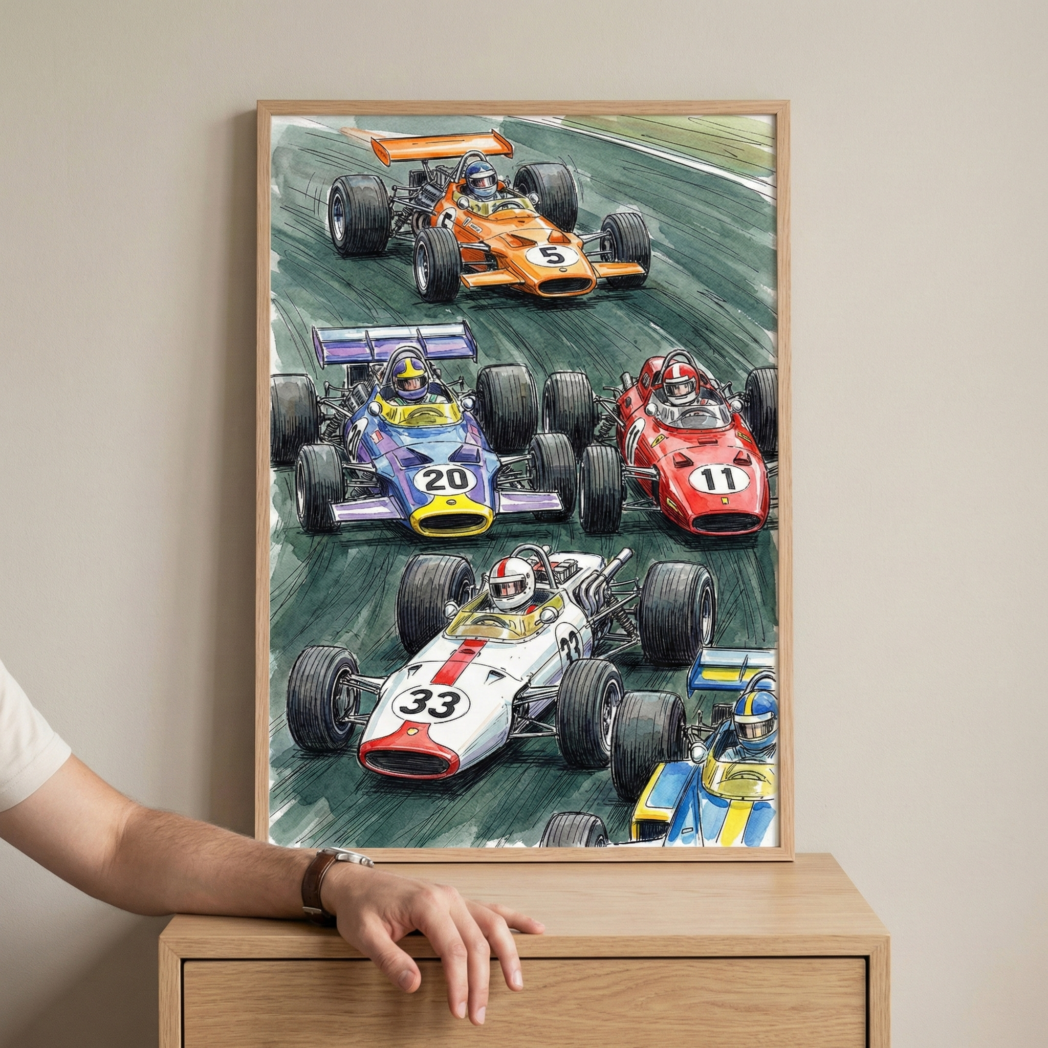 A framed illustration shows five vintage-style open-wheel race cars in a dynamic scene on a track. The cars, in vibrant colors like orange, purple, red, and white, feature large numbers such as 5, 20, 11, and 33. The artwork uses bold lines and color washes to suggest high speed. The frame rests on a wooden surface, with a person's arm and hand visible beside it.
