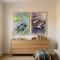 A bright room features two framed illustrations above a light oak dresser. The left image depicts a blue vintage racing car speeding around a track curve. The right artwork shows an orange and a purple racing car side-by-side, with dynamic motion lines conveying high speed. On the dresser sits a brass desk lamp, three wooden toy cars, and a stack of colorful books. A corner of a bed with a vibrant checkered wool blanket is visible in the foreground, set against neutral cream walls and a woven rug.