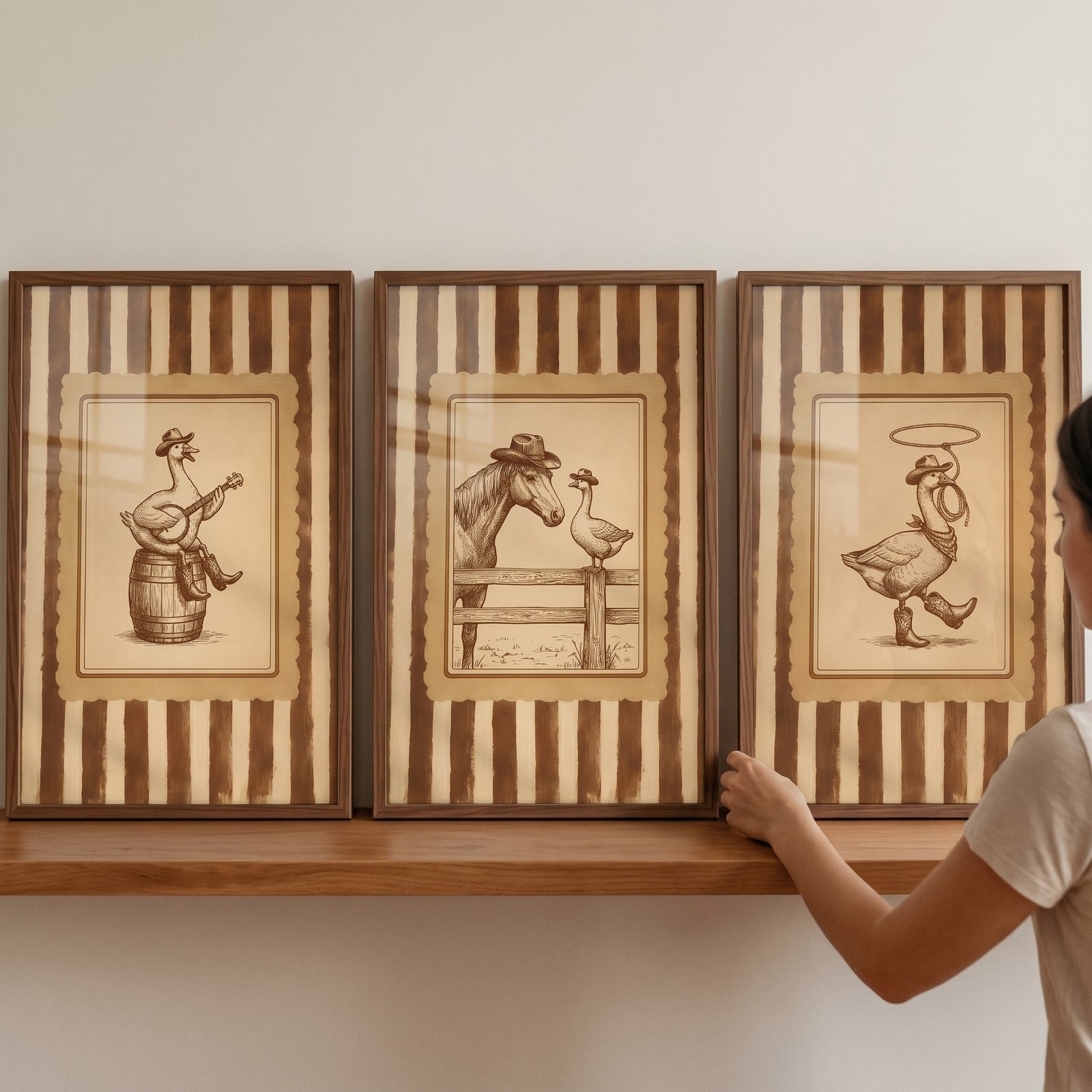 A person's hand adjusts a vintage sepia cowboy goose wall art set of 3 on a shelf, featuring a banjo, horse, and lasso.