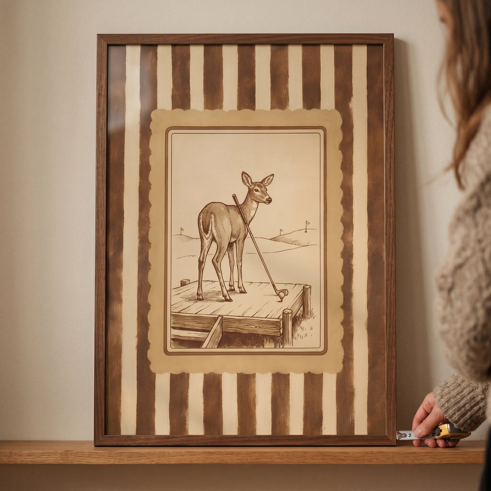 Vintage sepia golfing deer wall art in a wood frame with a striped border being measured with a tape measure on a shelf.
