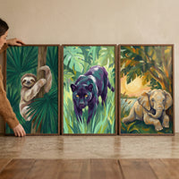 Three framed jungle animal paintings lean against a wall on a wooden floor while a person adjusts the left frame. The artworks show a smiling sloth hanging from a tree, a black panther walking through green foliage, and a baby elephant resting near water at sunset.