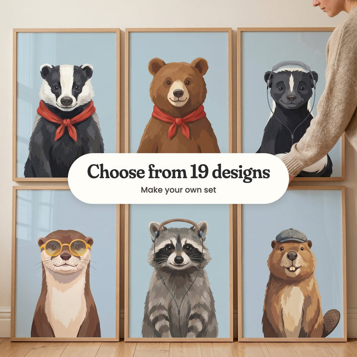 Six animal portrait prints in light wood frames in two rows leaning against a cream wall on a wood floor, with a woman's arm in a knit sweater setting one in place: a badger in a red neckerchief, a brown bear in a red bandana, a skunk in grey headphones, an otter in yellow round glasses, a raccoon in headphones and a beaver in a grey newsboy cap, with a label reading Choose from 19 designs, Make your own set