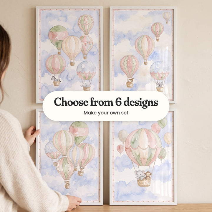 A hand straightening one of four hot air balloon prints in white frames in two rows on a cream wall: six balloons carrying a zebra and an elephant, five balloons with a fawn and a koala, a cluster of balloons with a teddy bear and a fawn, and one big balloon carrying an elephant and a monkey, with a label reading Choose from 6 designs, Make your own set