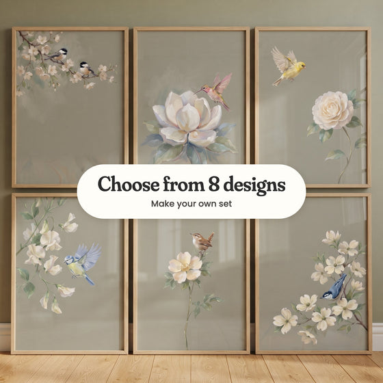 Six songbird prints in light wood frames leaning on the floor in two rows against a sage wall: two chickadees on white blossom, a hummingbird at a magnolia, a yellow finch above a cream bloom, a blue tit by sweet peas, a wren on a wild rose and a nuthatch in dogwood, with a label reading Choose from 8 designs, Make your own set