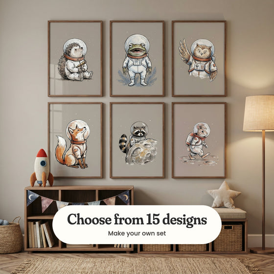Taupe kids' room with six astronaut animal prints in dark wood frames in two rows above a wood bookshelf hung with moon and star bunting and a cushioned bench with woven baskets, beside a plush rocket and a floor lamp: Spiky Space Explorer, Cosmic Croaker, Cosmic Owl Voyager, Stellar Space Fox, Lunar Space Bandit and Lunar Rabbit Walk, with a label reading Choose from 15 designs, Make your own set