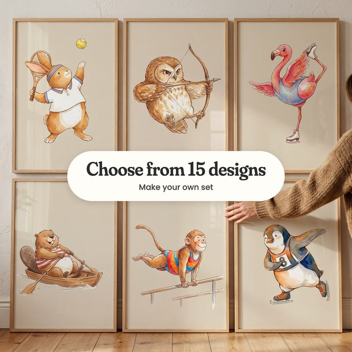 Six sporty animal prints in light wood frames leaning on the floor in two rows: a bunny serving at tennis, an owl with a bow and arrow, a flamingo ice skating, a beaver rowing, a monkey on parallel bars and a penguin speed skating, with a label reading Choose from 15 designs, Make your own set