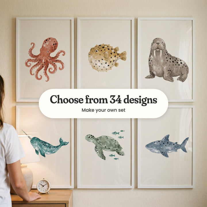 Six spotted sea animal prints in white frames in two rows on a cream wall, beside a woman at a wood dresser with a lamp and an alarm clock: a rust octopus, a mustard pufferfish, a walrus, a teal narwhal, a sea turtle swimming with small fish and a grey-blue shark, with a label reading Choose from 34 designs, Make your own set