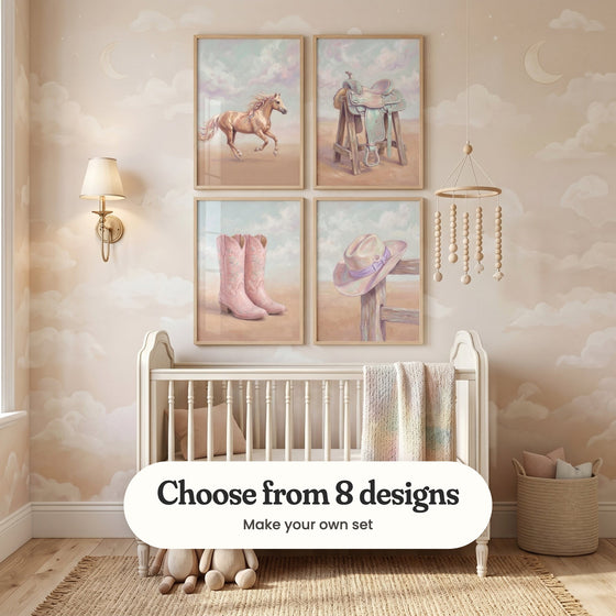 Blush nursery with cloud and moon wallpaper and four western prints in light wood frames in two rows above a white crib with a knit blanket, beside a brass wall lamp, a bead mobile, a woven basket and plush animals: Pastel Ribbon Gallop, Pastel Desert Dream, Blossom Western Boots and Lavender Ranch Hat, with a label reading Choose from 8 designs, Make your own set
