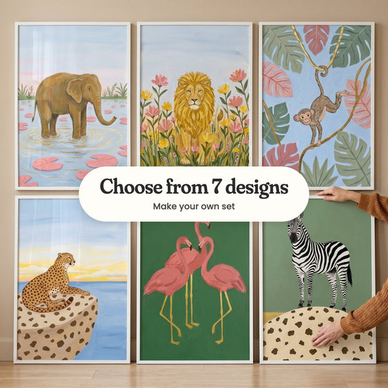 Six painterly safari animal prints in white frames leaning in two rows on a wood floor, with hands in rust-orange knit sleeves: an elephant in a lily pond, a lion among flowers, a monkey swinging on golden vines, a cheetah on a spotted rock by the sea, three pink flamingos and a zebra on sage green, with a label reading Choose from 7 designs, Make your own set