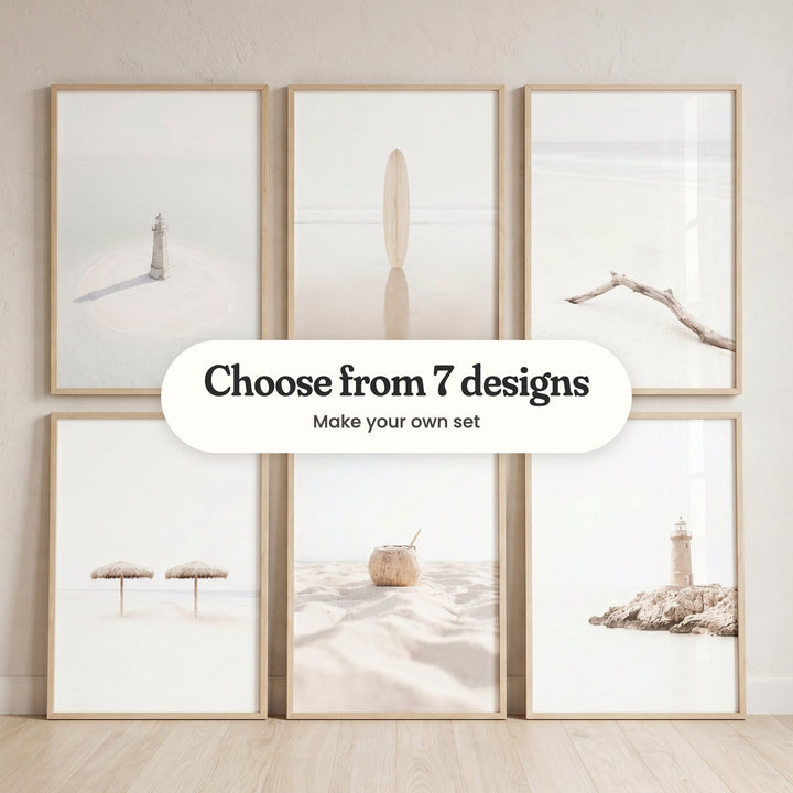 Six beach prints in light wood frames against a cream wall, three hanging on the wall above three leaning on a pale wood floor: a small white lighthouse on a sandbar, a wooden surfboard standing in shallow water, a bleached driftwood branch on the sand, two straw umbrellas on white sand, a coconut with a straw and a lighthouse on a rocky outcrop, with a label reading Choose from 7 designs, Make your own set