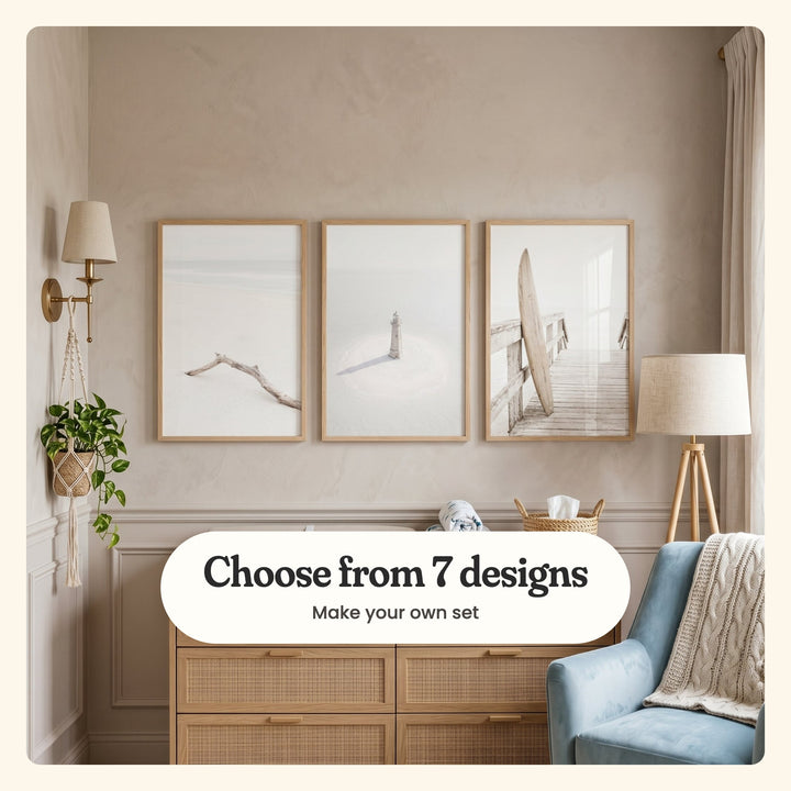 Nursery with three beach prints in light wood frames above a rattan-front dresser with a woven basket and a rolled muslin, beside a brass wall sconce, a macrame plant hanger, a tripod floor lamp and a blue velvet armchair with a cable-knit throw: a bleached driftwood branch on the sand, a small white lighthouse on a sandbar and a wooden surfboard leaning on a boardwalk railing, labeled Choose from 7 designs, Make your own set