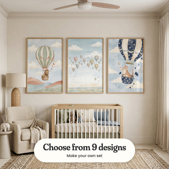 Warm cream nursery with three balloon prints in light wood frames above a light wood crib with plush teddy bears and sage and blue cushions, beside a rattan floor lamp, a cream armchair with a knit throw, a small elephant figure and a fringed rug: Lion Sky Voyage, Whimsical Sky Voyage and Starlight Tiger Voyage, with a label reading Choose from 9 designs, Make your own set