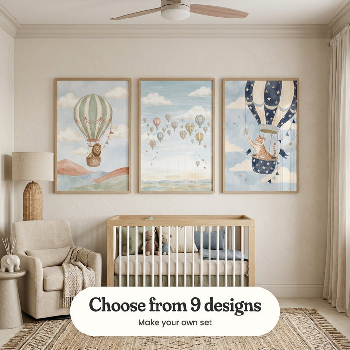 Warm cream nursery with three balloon prints in light wood frames above a light wood crib with plush teddy bears and sage and blue cushions, beside a rattan floor lamp, a cream armchair with a knit throw, a small elephant figure and a fringed rug: Lion Sky Voyage, Whimsical Sky Voyage and Starlight Tiger Voyage, with a label reading Choose from 9 designs, Make your own set