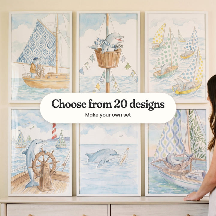 Six dolphin sailor prints in white frames standing on a cream dresser, with a woman beside them: a dolphin captain at the wheel, a dolphin in a crow's nest, dolphins in polka-dot sailboats, a dolphin at a carved wheel with a lighthouse, a dolphin with a message in a bottle and a dolphin napping in a hammock, with a label reading Choose from 20 designs, Make your own set